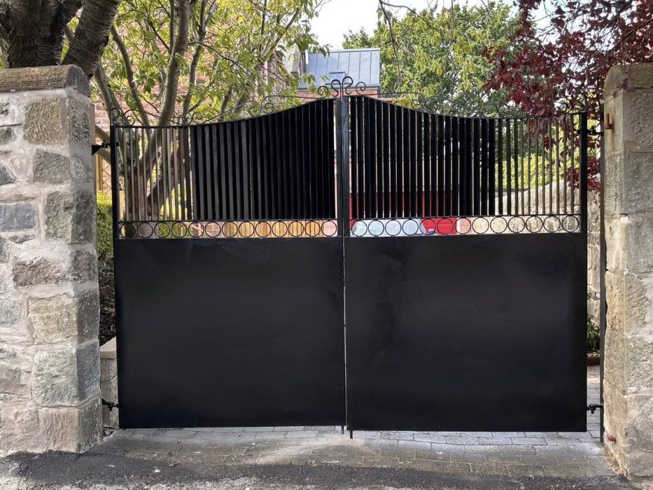 Black driveway gate with scrolled cresting and a band of forged circles, hung between stone piers