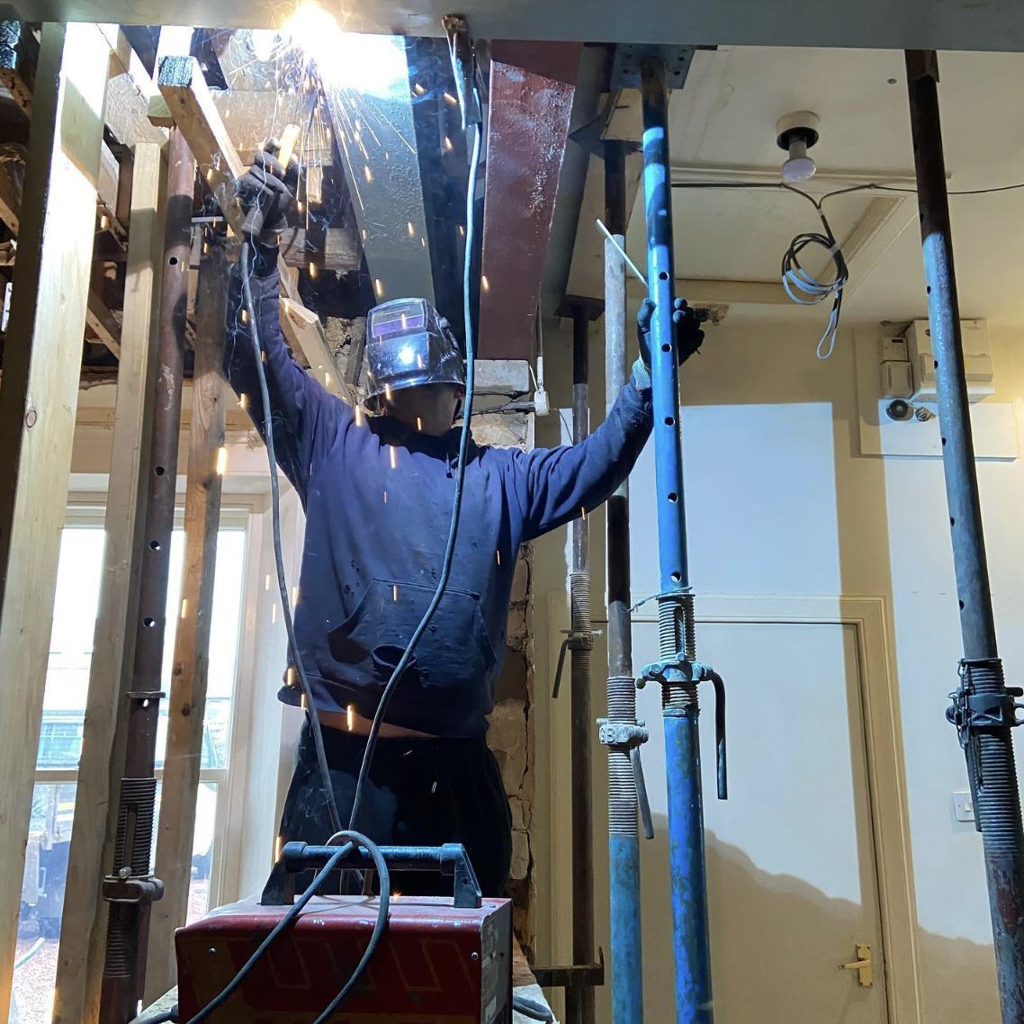 A welder working overhead between acro props, sparks falling, installing a steel beam inside a house